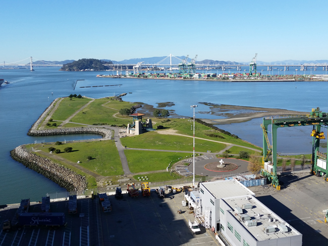 Aerial photo of the Port of Oakland at low tide. An industrial bulding on a concrete area is in the foreground with a green iron beam structure to the lower-right. A grassy penninsula extends into the water with criss-crossed roads. In the center right, water is over low land areas. In the upper portion is another structure resembling a very long barge or a man made penninsula with industrial constructions. A bridge and mountains are in the distance.